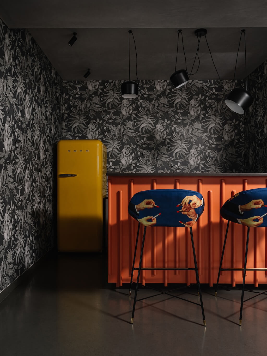 Bar area with a bold orange counter, floral wallpaper, yellow retro fridge, and quirky blue chairs, reflecting eclectic and vibrant design choices