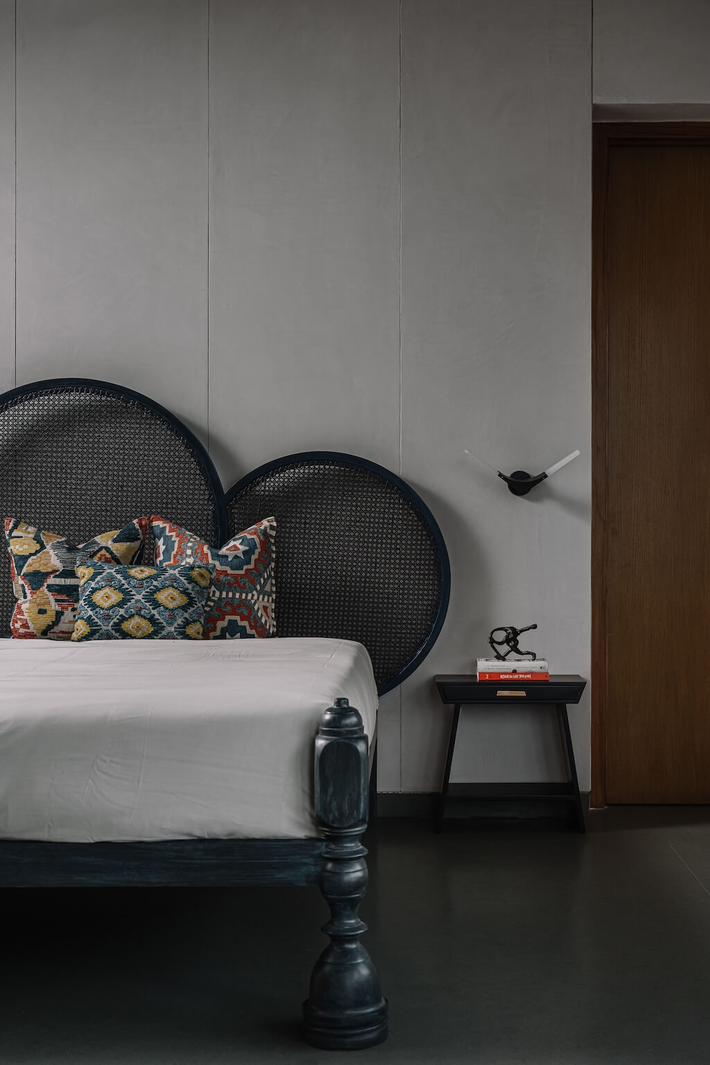 Bedroom with a dark rattan headboard, patterned cushions, minimalist side table with decor, and muted walls, creating a calm and personalized aesthetic
